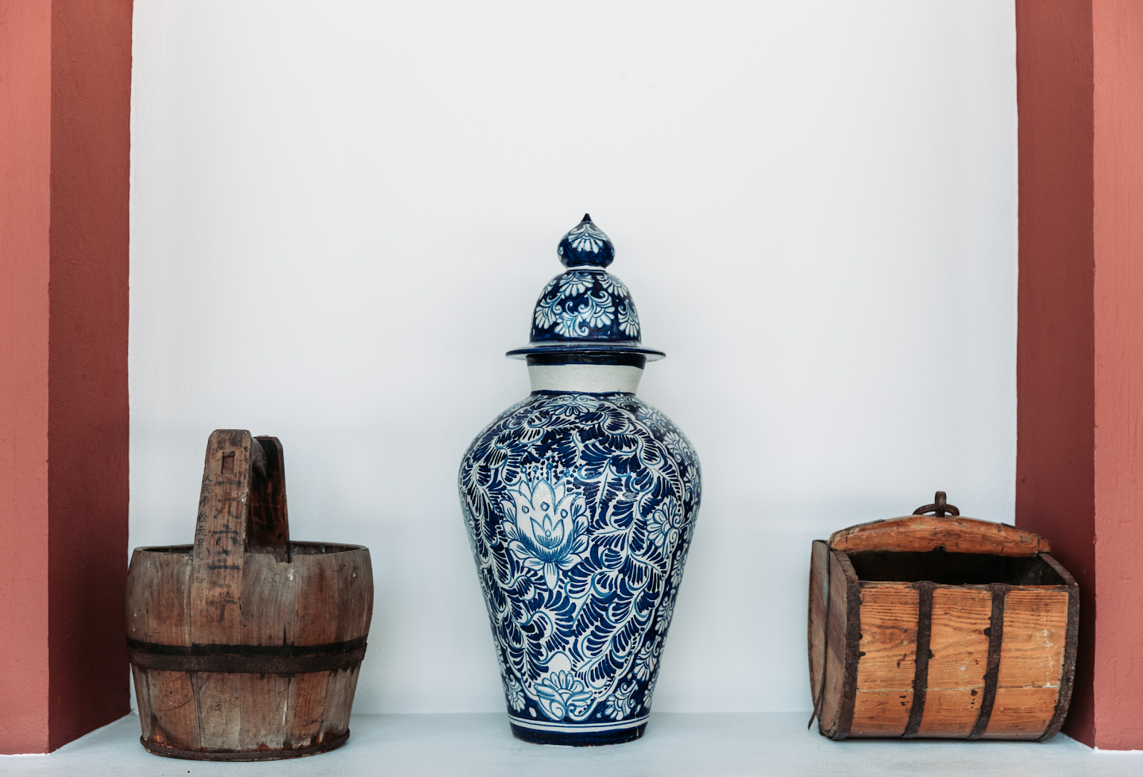 A tall blue-and-white painted jar with a domed lid standing in a whitewashed alcove, an old wooden bucket on either side of it.