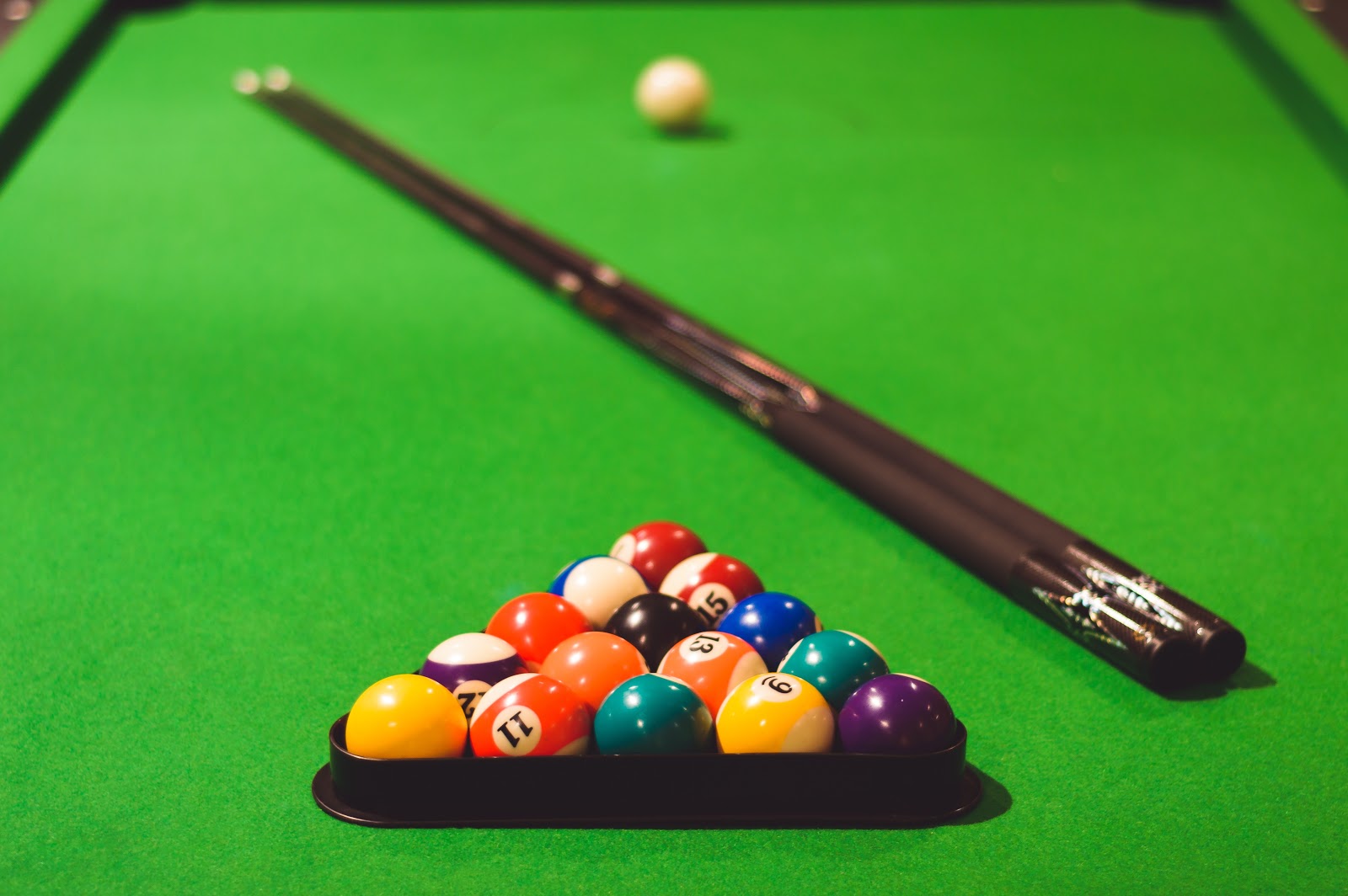 A rack of billiard balls waiting in the triangle on a green table, the cue laid across the baize behind them and the cue ball beyond.